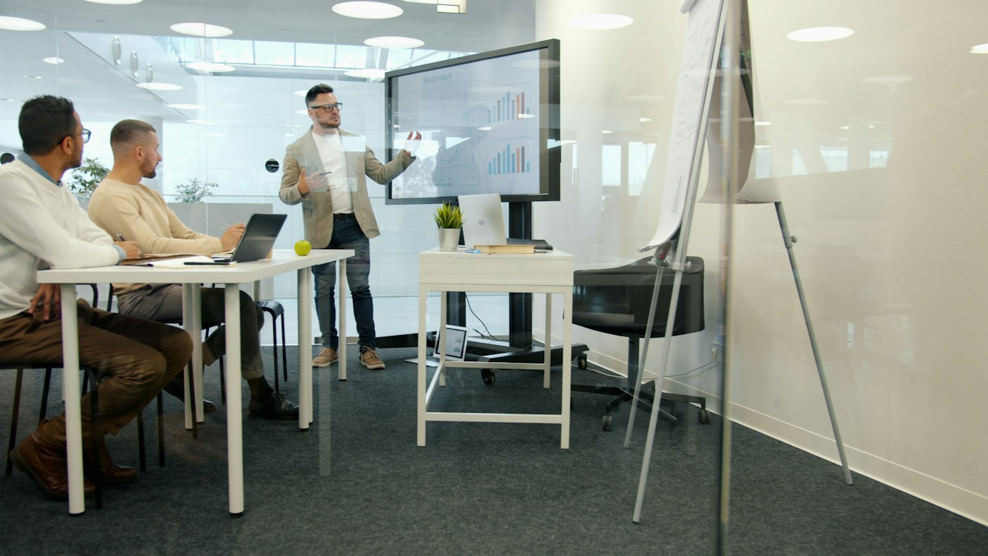 A professional presenting sales information beside a monitor in a boardroom